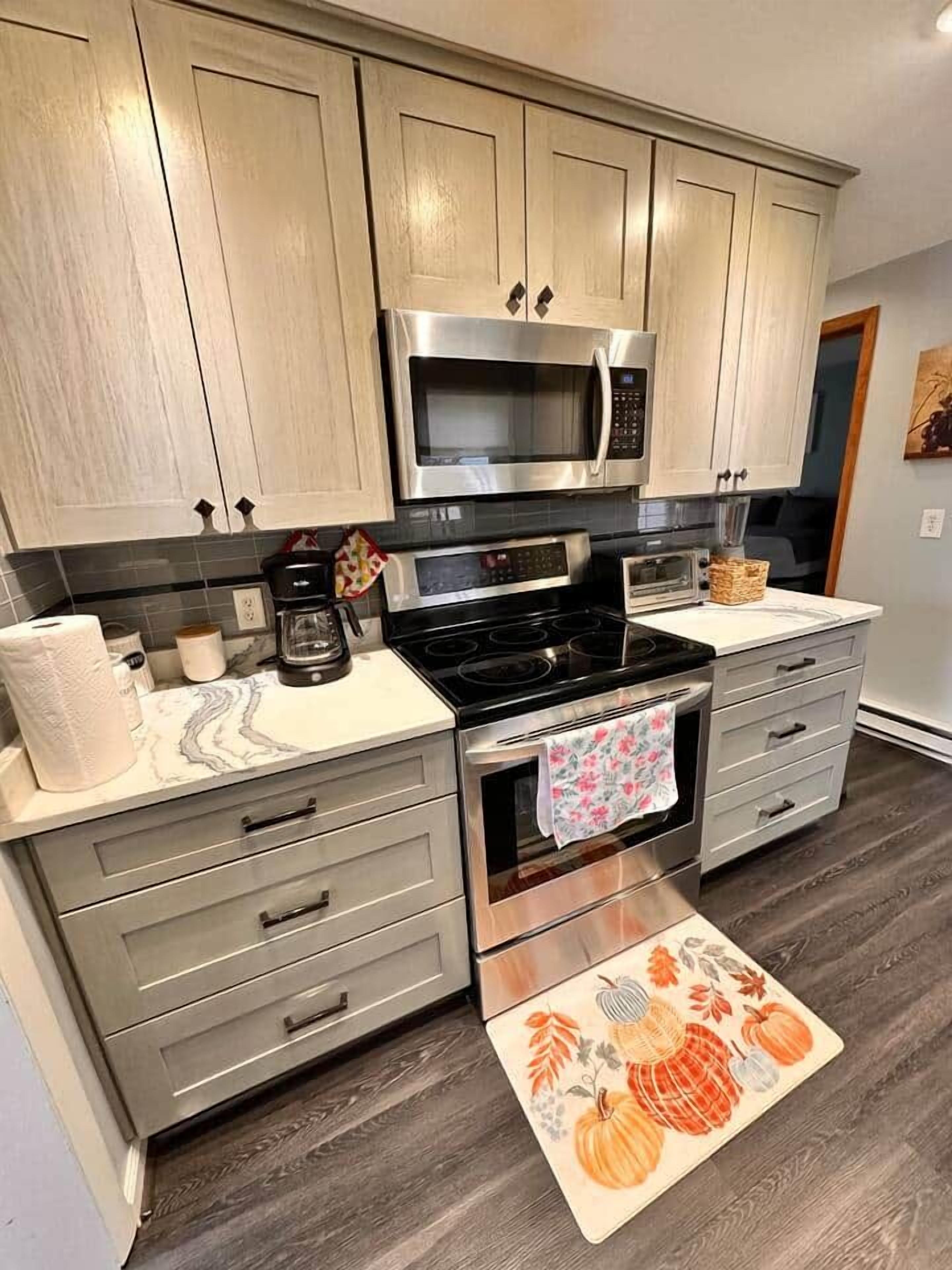 A kitchen with a microwave, stove, and oven, light wood cabinets, and a patterned rug.