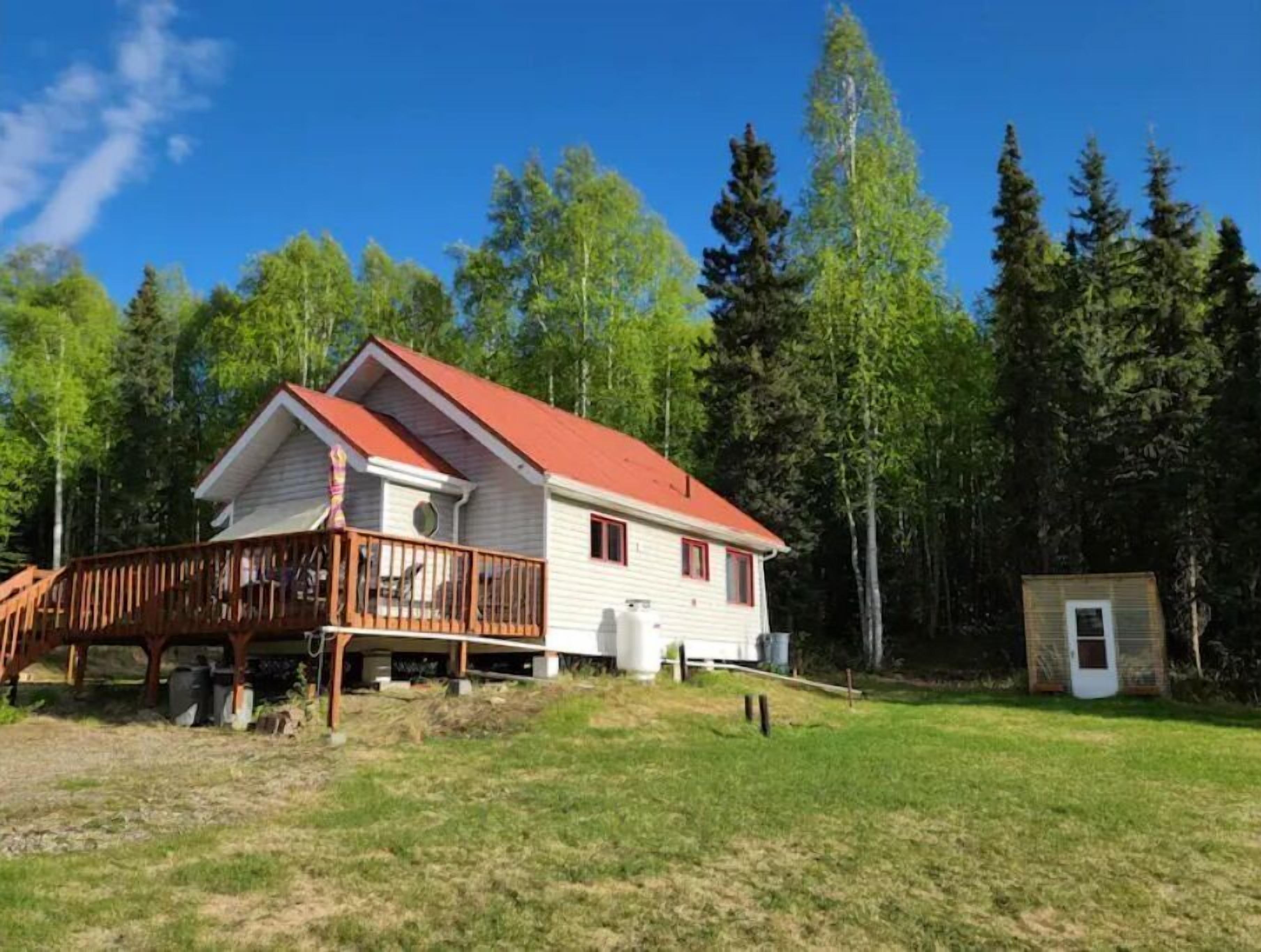 A two-story cabin with a red roof, a wooden deck, and a small shed in a forested area.