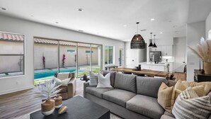 Ein modernes Wohnzimmer mit einem großen grauen Sofa, einem Holztisch und Blick auf einen Swimmingpool durch große Schiebeglastüren.