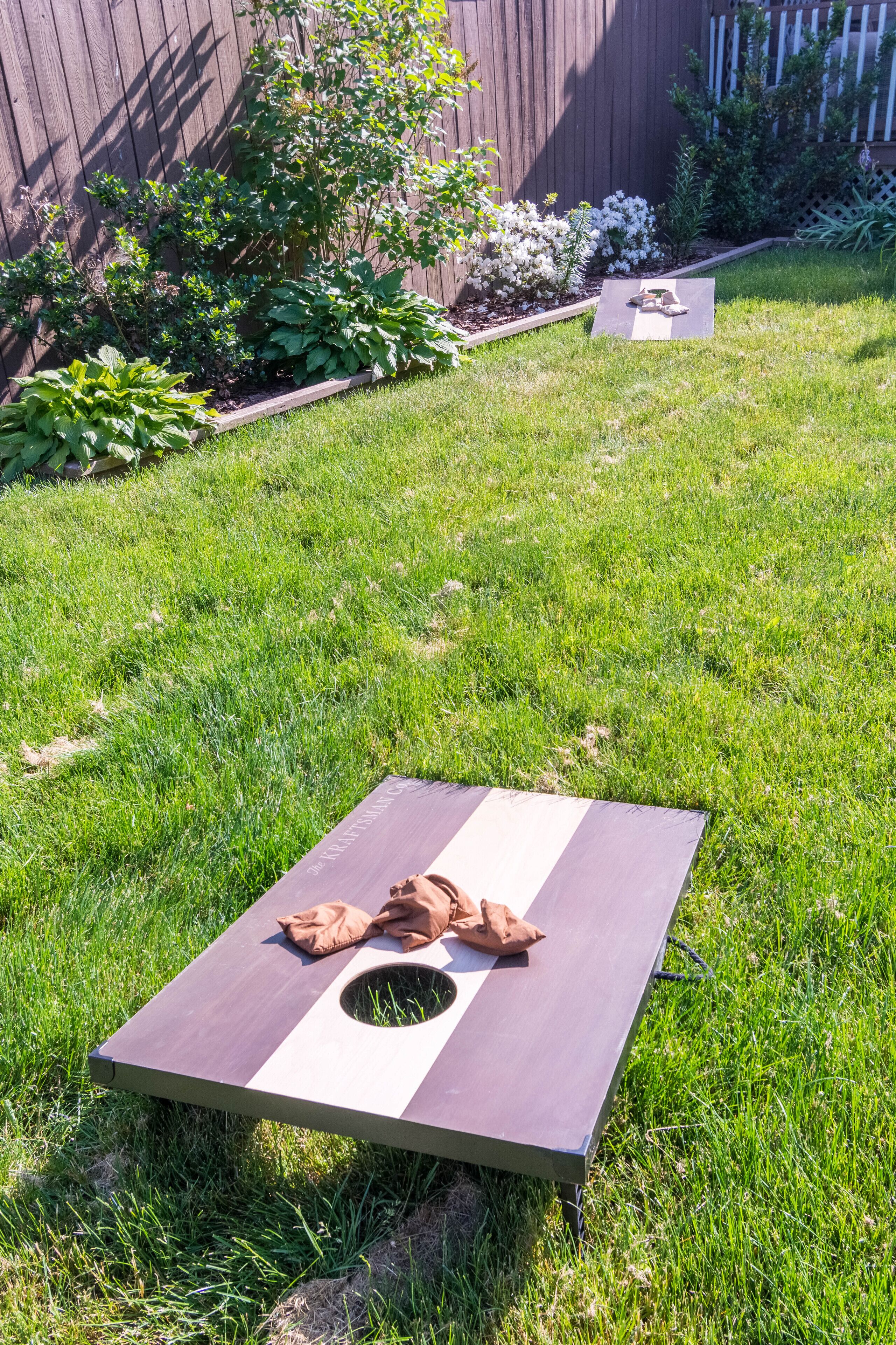 A large wooden board game set up on a grassy lawn with a wooden fence and flowering plants in the background.
