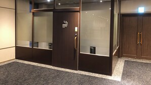A modern, enclosed smoking area with a dark wooden door, a glass partition, and a small trash bin. - SK HOTEL KOBE STATION FRONT (Kobe)