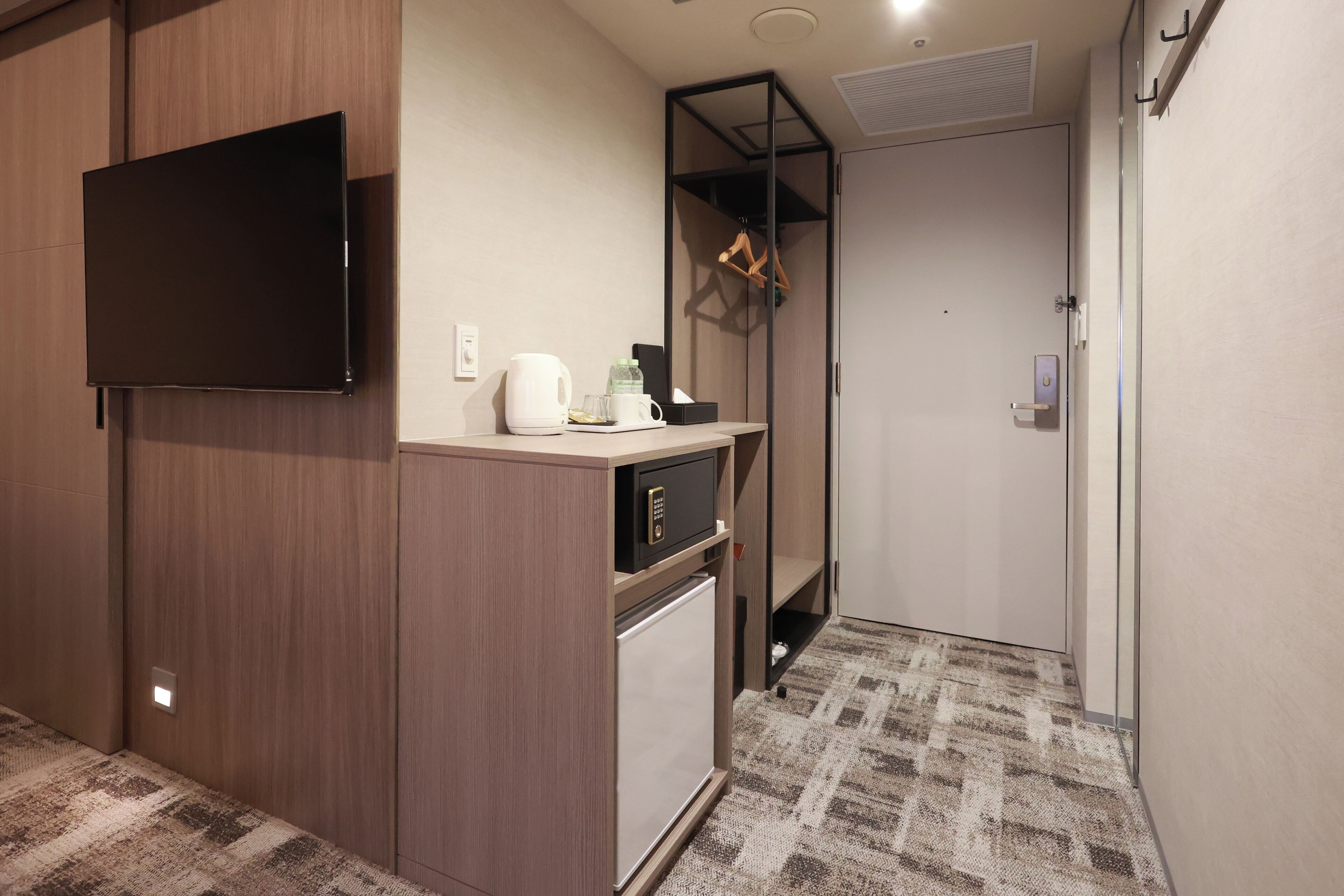 Habitación de hotel compacta con televisión de pantalla plana, minibar, refrigerador pequeño, cafetera y perchero.