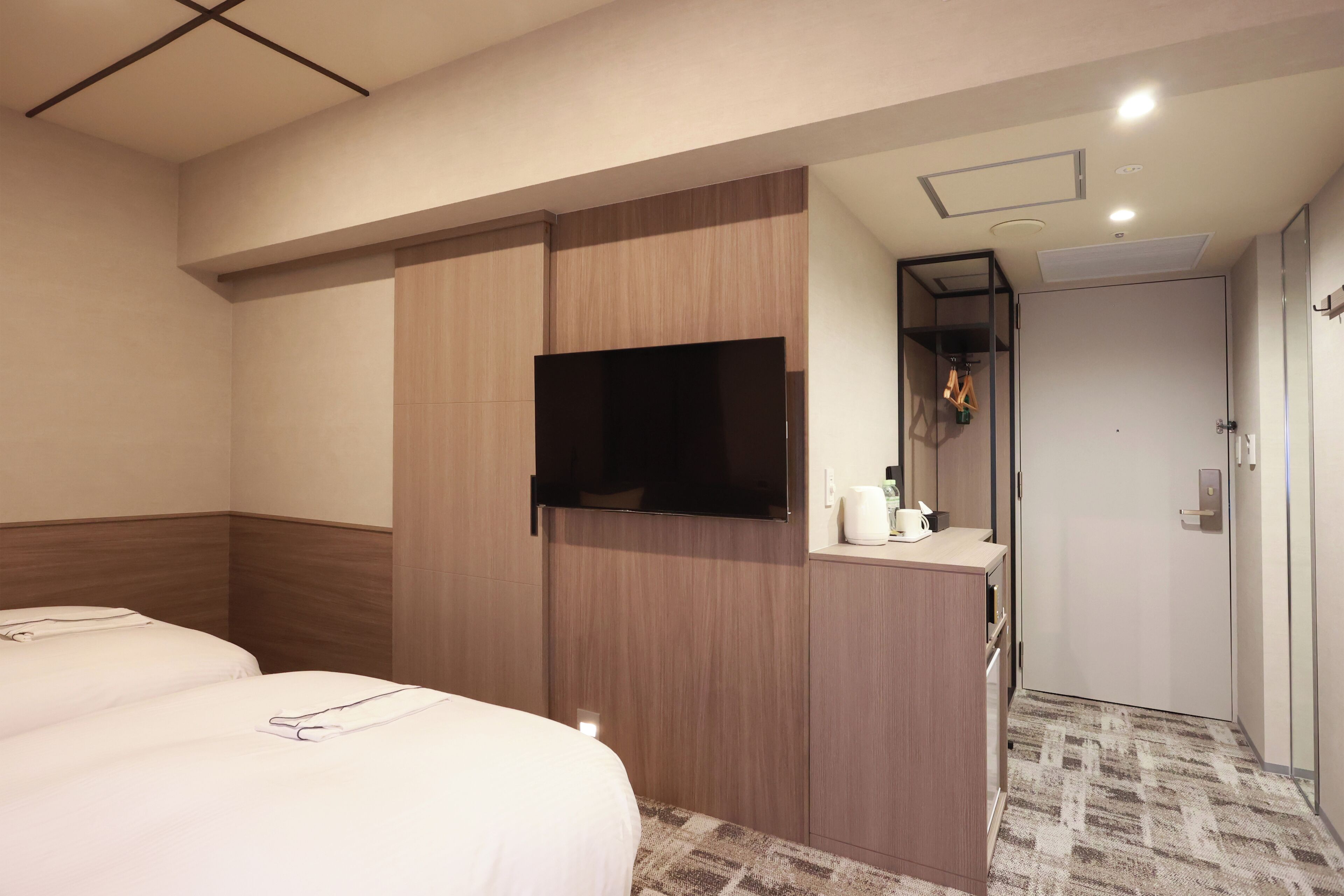 A hotel room with two beds, a TV mounted on a wooden cabinet, a small desk with a mirror, and a door leading to a bathroom.