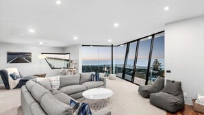 Uma sala de estar moderna com um amplo sofá modular, uma mesa de centro redonda e vista para o oceano através de janelas do chão ao teto.