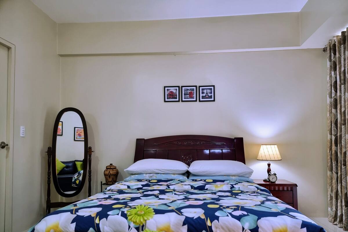 A neatly made bed with a floral bedspread, a bedside lamp, and a clock on the nightstand.