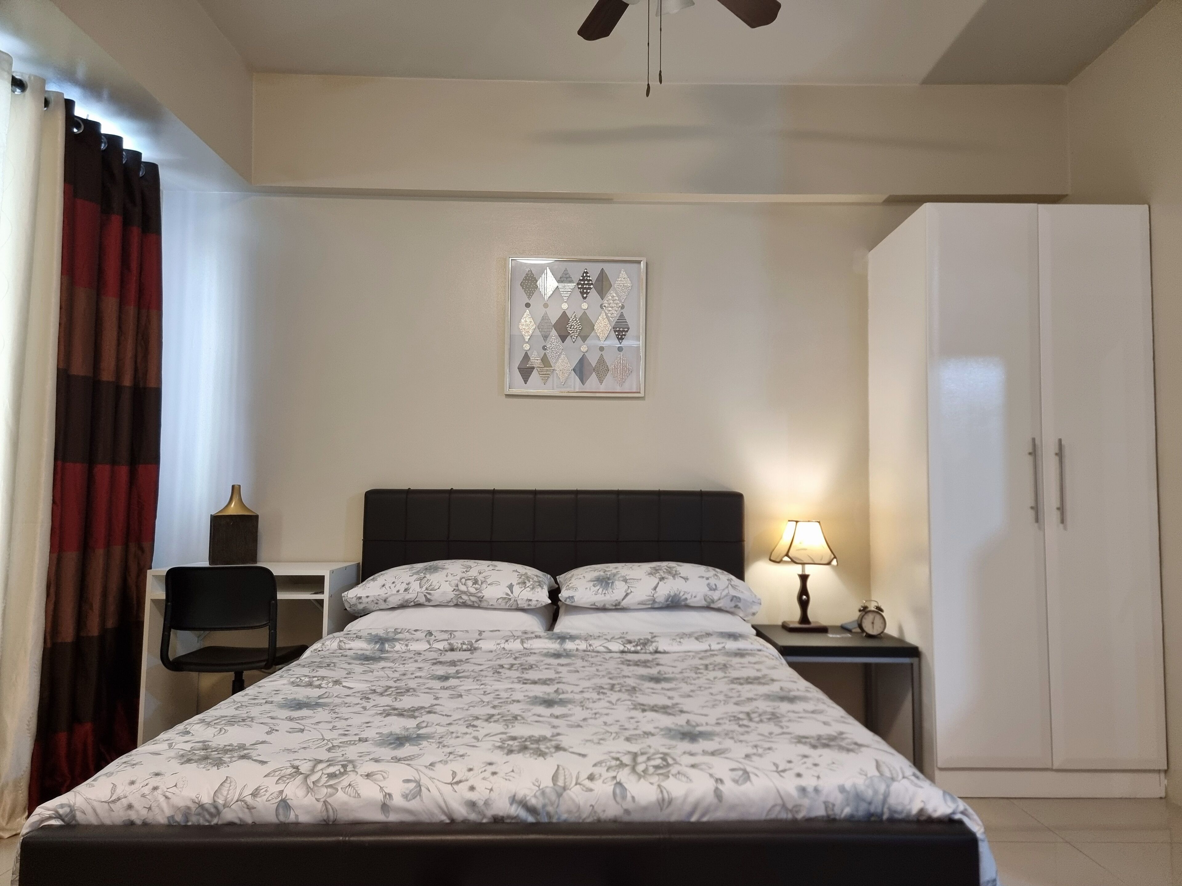 A bedroom with a bed, a nightstand, a lamp, a chair, a wardrobe, and a ceiling fan.