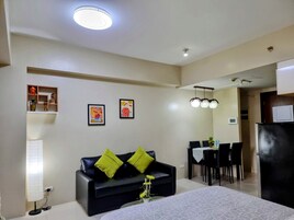 A modern hotel room with a black leather sofa, a bed with a patterned bedspread, a dining area with a table and chairs, and a kitchenette with a refrigerator.