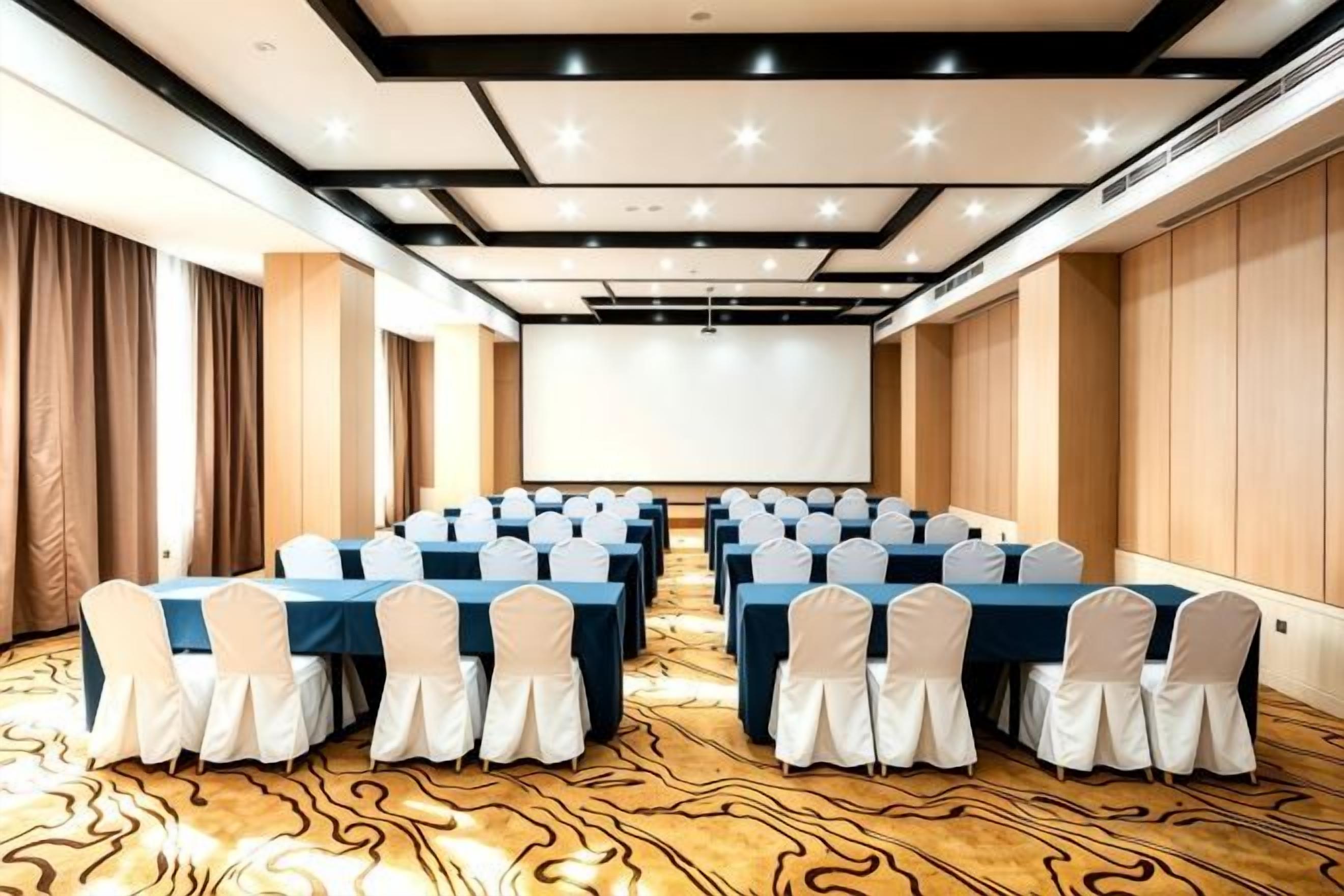 A conference room with rows of tables and chairs, a projector screen, and a patterned carpet.