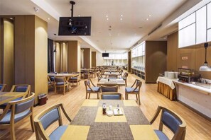 A spacious restaurant with multiple dining tables and chairs, a television mounted on the ceiling, and a kitchen area visible on the right. - Xierdun Yixuan Hotel (Lishui)