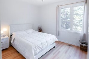 A bedroom with a bed, a nightstand, a lamp, a window with a view of trees, and a basket on the floor.