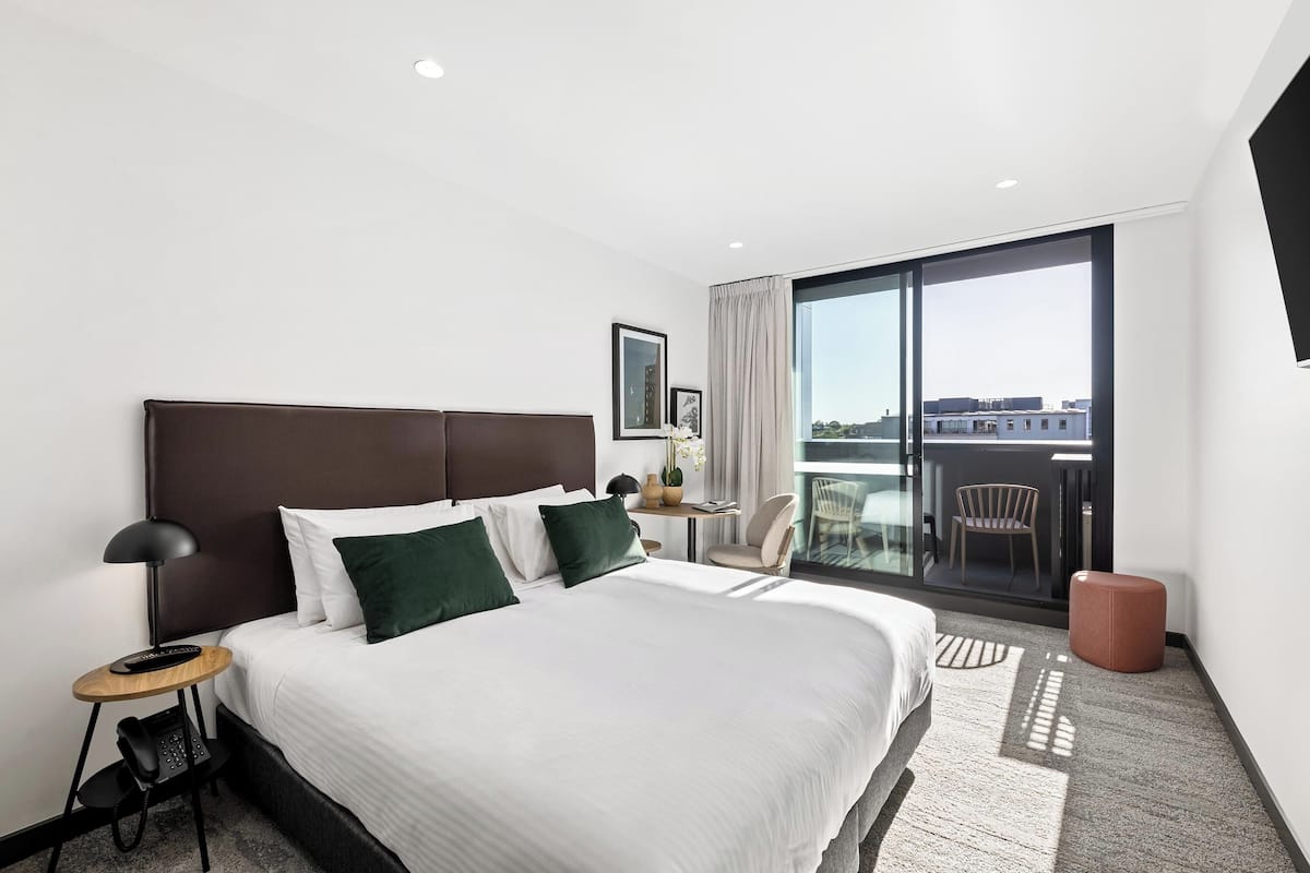 A modern hotel room with a large bed, a desk with a lamp, a chair, and a view of the city through large windows.