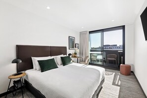 A modern hotel room with a large bed, a desk with a lamp, a chair, and a view of the city through large windows. - Quest Geelong Central (Geelong)