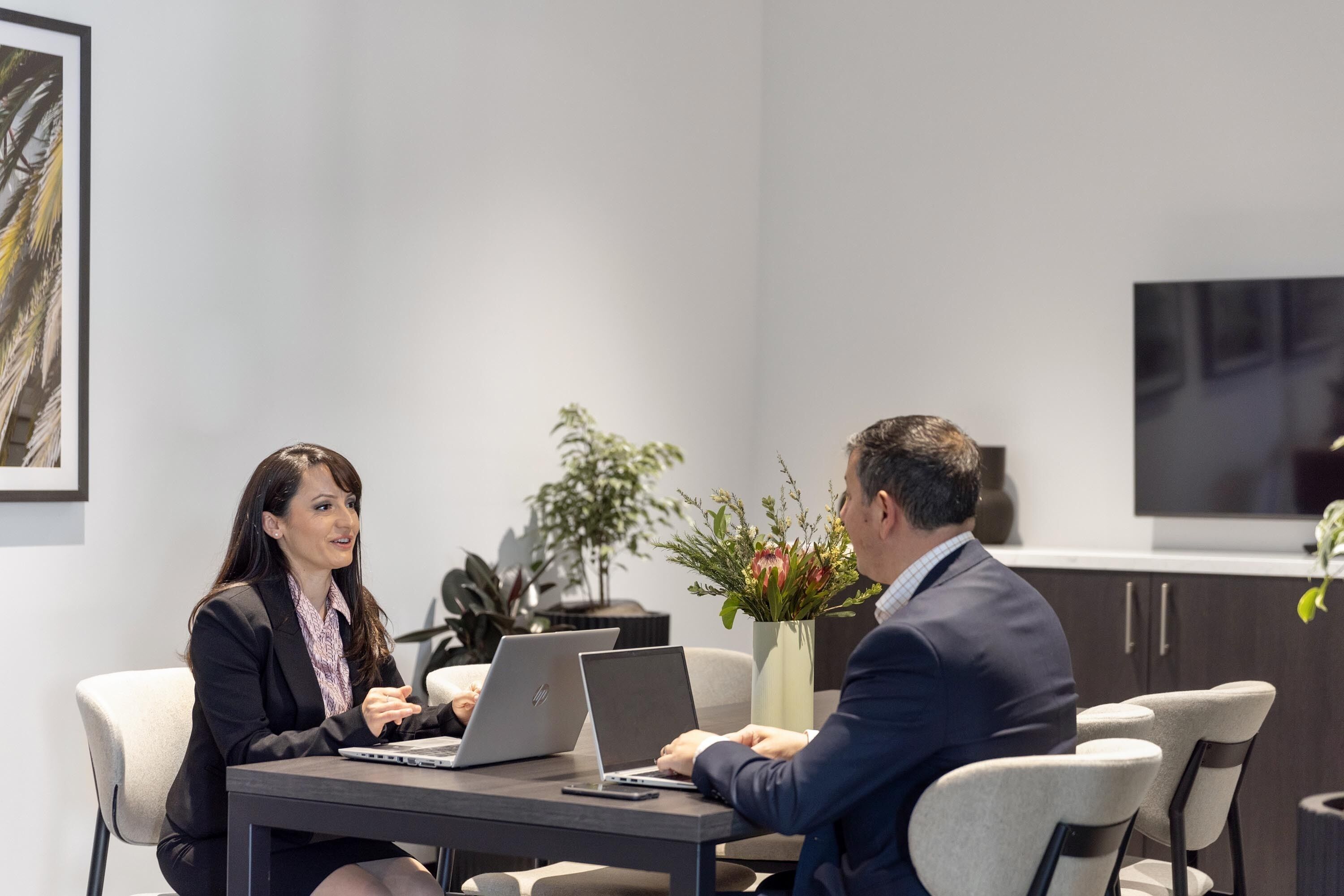 A modern office setting with two individuals at a table with laptops, surrounded by plants and a large screen.