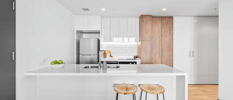 A modern kitchen with white cabinetry, a wooden accent panel, a stainless steel refrigerator, and two wooden stools.