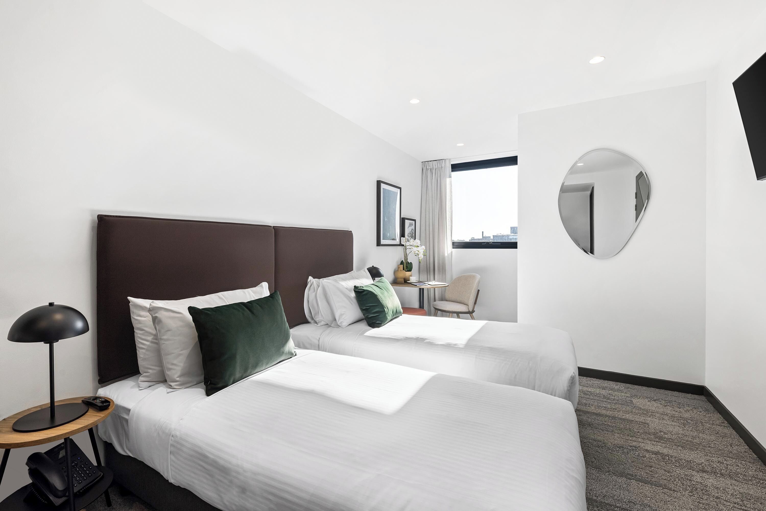 A hotel room with two beds, a desk, a chair, a round mirror, and a window with a city view.