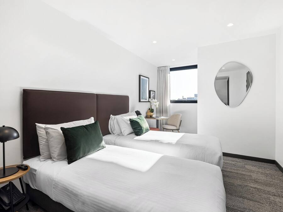 Habitación de hotel con dos camas, un escritorio, una silla, un espejo redondo y una ventana con vista a la ciudad.