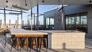 Un área de bar moderna al aire libre con una barra de concreto, taburetes de metal y amplios ventanales con vistas a la ciudad.