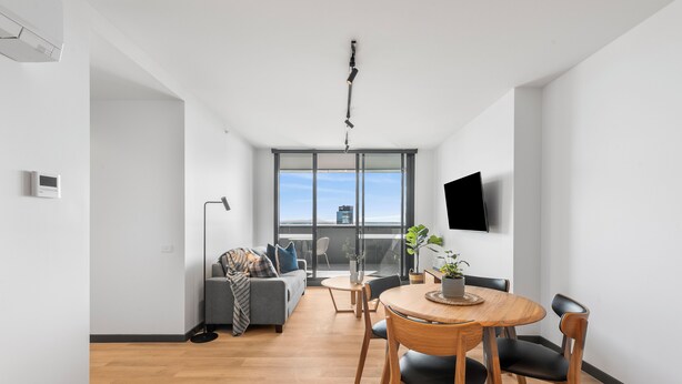 Un moderno salón con sofá, mesa de comedor y sillas. Hay un televisor montado en la pared y una ventana con vista a la ciudad.