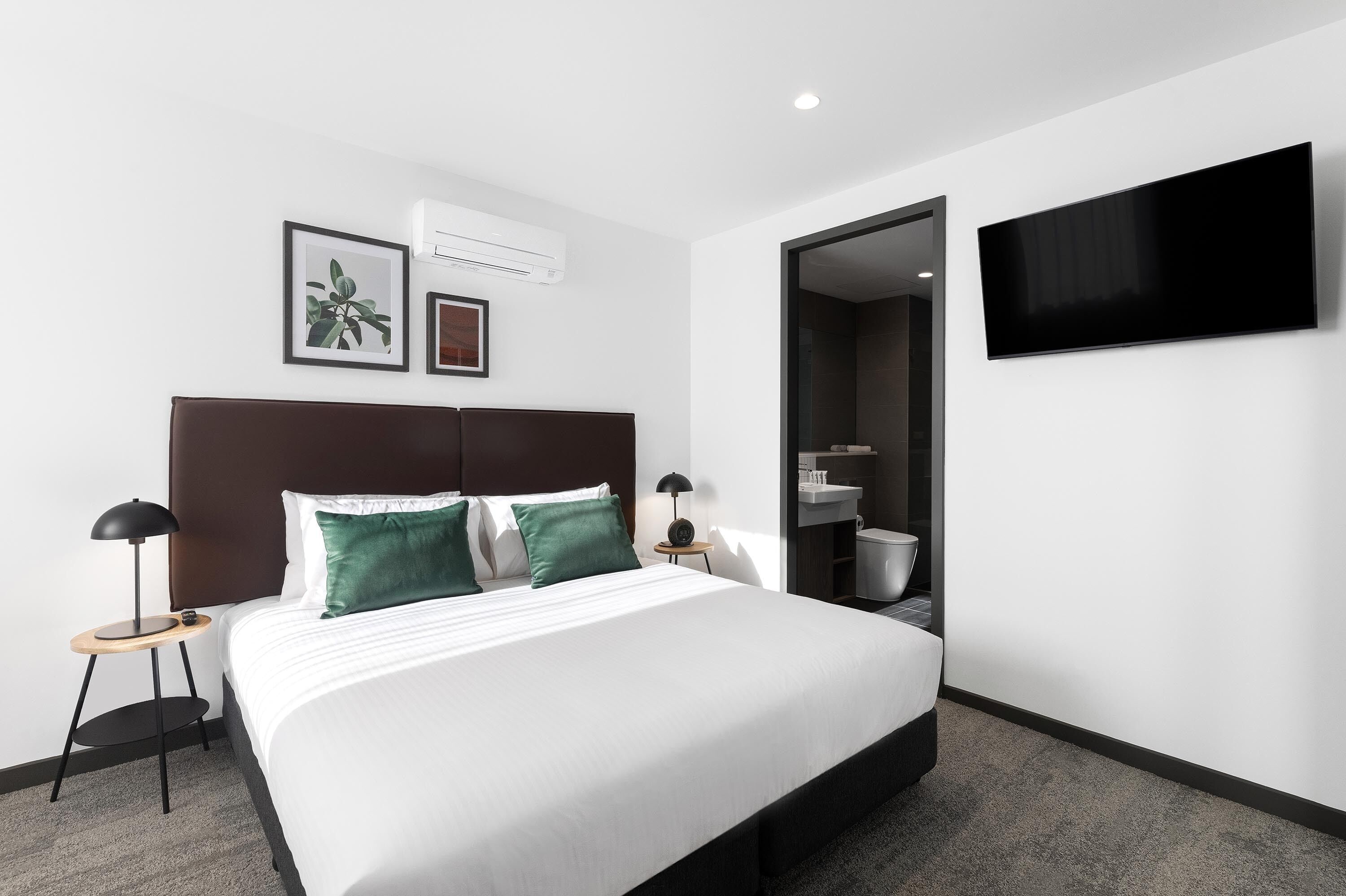 A modern hotel room with a large bed, two bedside tables with lamps, a flat-screen TV mounted on the wall, and a bathroom visible through an open door.