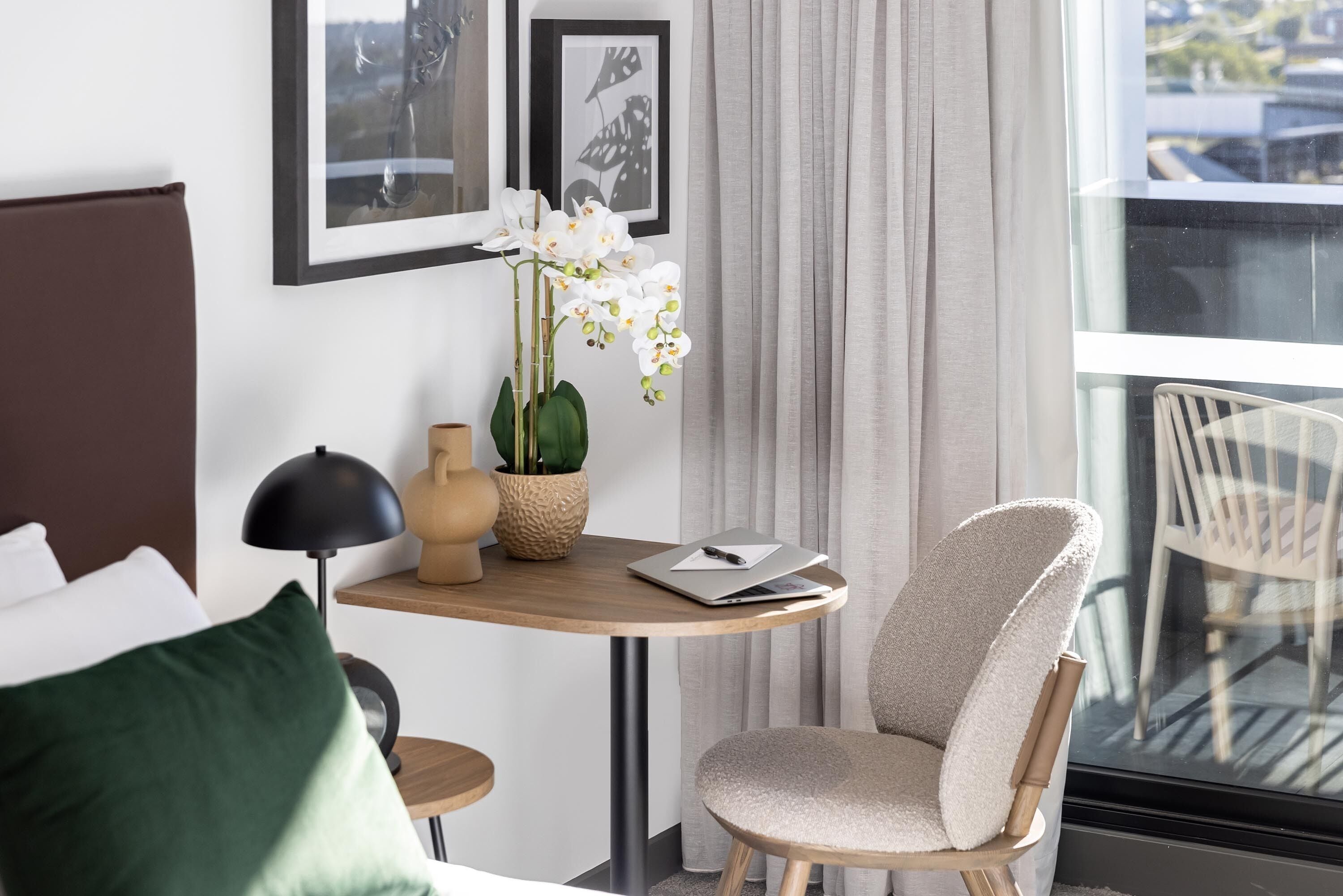 A modern hotel room with a bed, a small round table with a laptop and a vase of flowers, a chair, and a view of the outside through a large window.