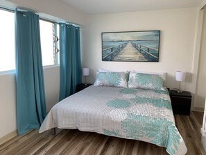 A bedroom with a bed, teal curtains, a painting of a pier, and a nightstand with a lamp. - Breezy Waikiki Apartment sleeps 6 with Free Parking (Honolulu)