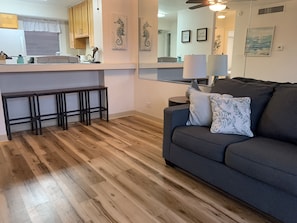 Un salon moderne avec un canapé gris, un plancher en bois et un coin cuisine doté de meubles en bois et d’un comptoir blanc.