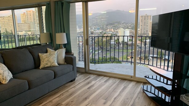 Ein modernes Wohnzimmer mit einem grauen Sofa, einem Fernseher und einem Balkon mit Blick auf die Stadt.
