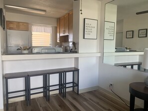 Une cuisine compacte avec des meubles en bois, un four encastré et une fenêtre avec des stores. On y trouve deux tabourets noirs et une petite table.