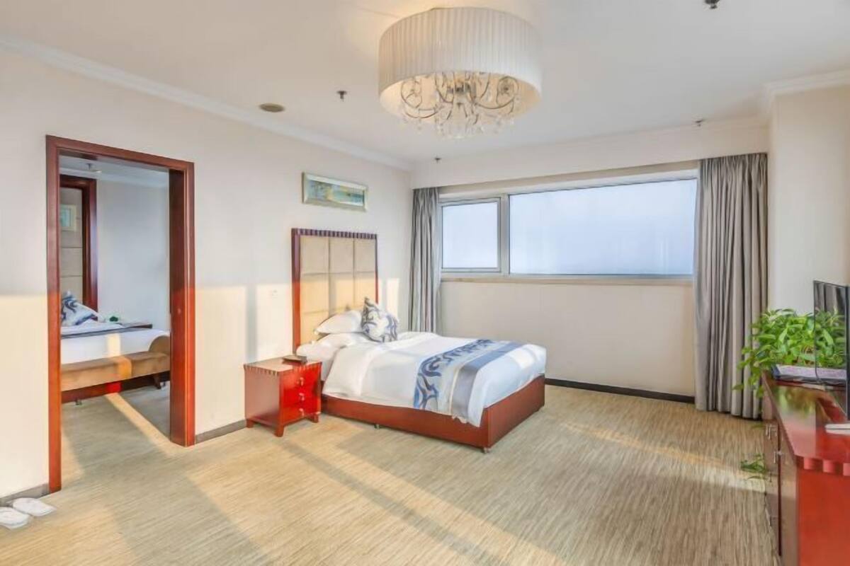 A hotel room with a large bed, a bedside table, a mirror, a window with curtains, and a chandelier.