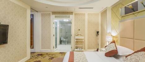 A hotel room with a bed, a television, a chandelier, and a bathroom visible through a glass door.