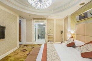 Habitación de hotel con cama, televisión, candelabro y baño visible a través de una puerta de cristal.