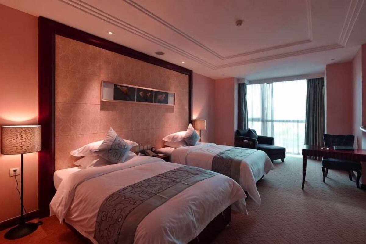 A hotel room with two beds, a desk, a chair, a lamp, and a large window.