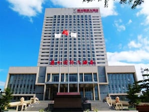 A multi-story building with a prominent entrance, a flagpole with flags, and a statue in front.