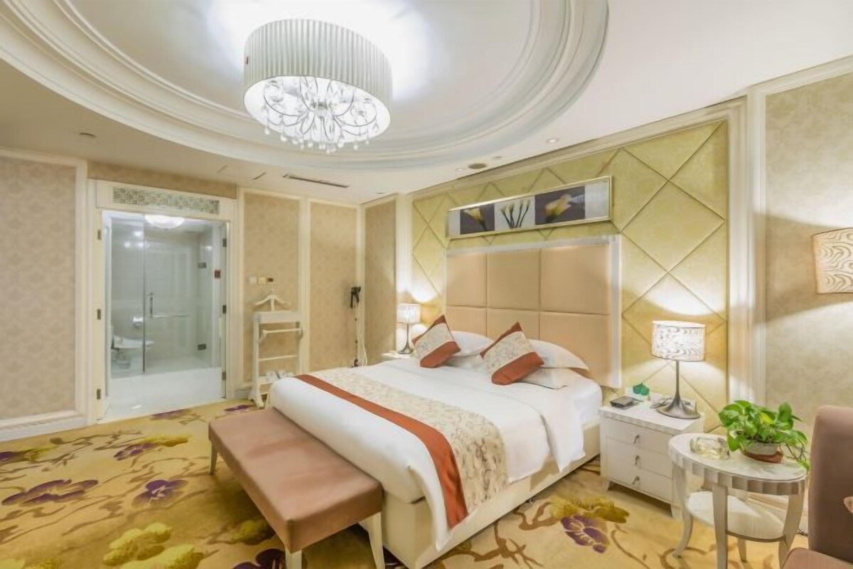 A hotel room with a bed, bedside table, lamp, chair, bench, and a door leading to a bathroom.