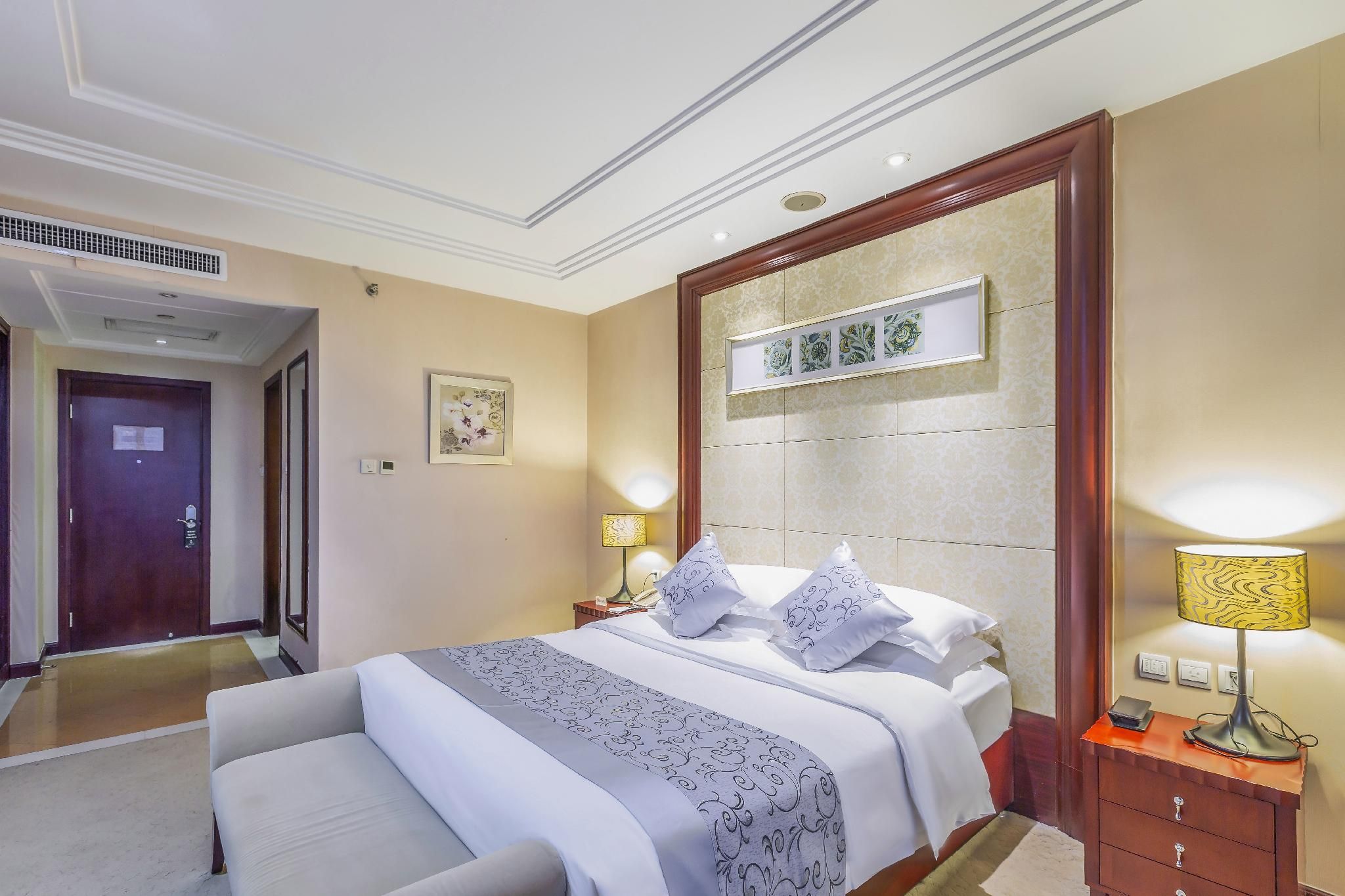 Habitación de hotel con una cama grande, lámparas de noche, una mesita de noche, una obra de arte enmarcada sobre la cama y una puerta que conduce a otra habitación.