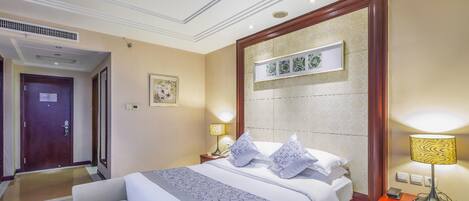 Habitación de hotel con una cama grande, lámparas de noche, una mesita de noche, una obra de arte enmarcada sobre la cama y una puerta que conduce a otra habitación.
