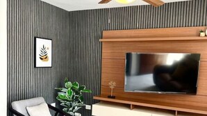 Una sala de estar moderna con una televisión de pantalla plana montada en una pared de madera, un ventilador de techo, un sillón gris y una planta en maceta.