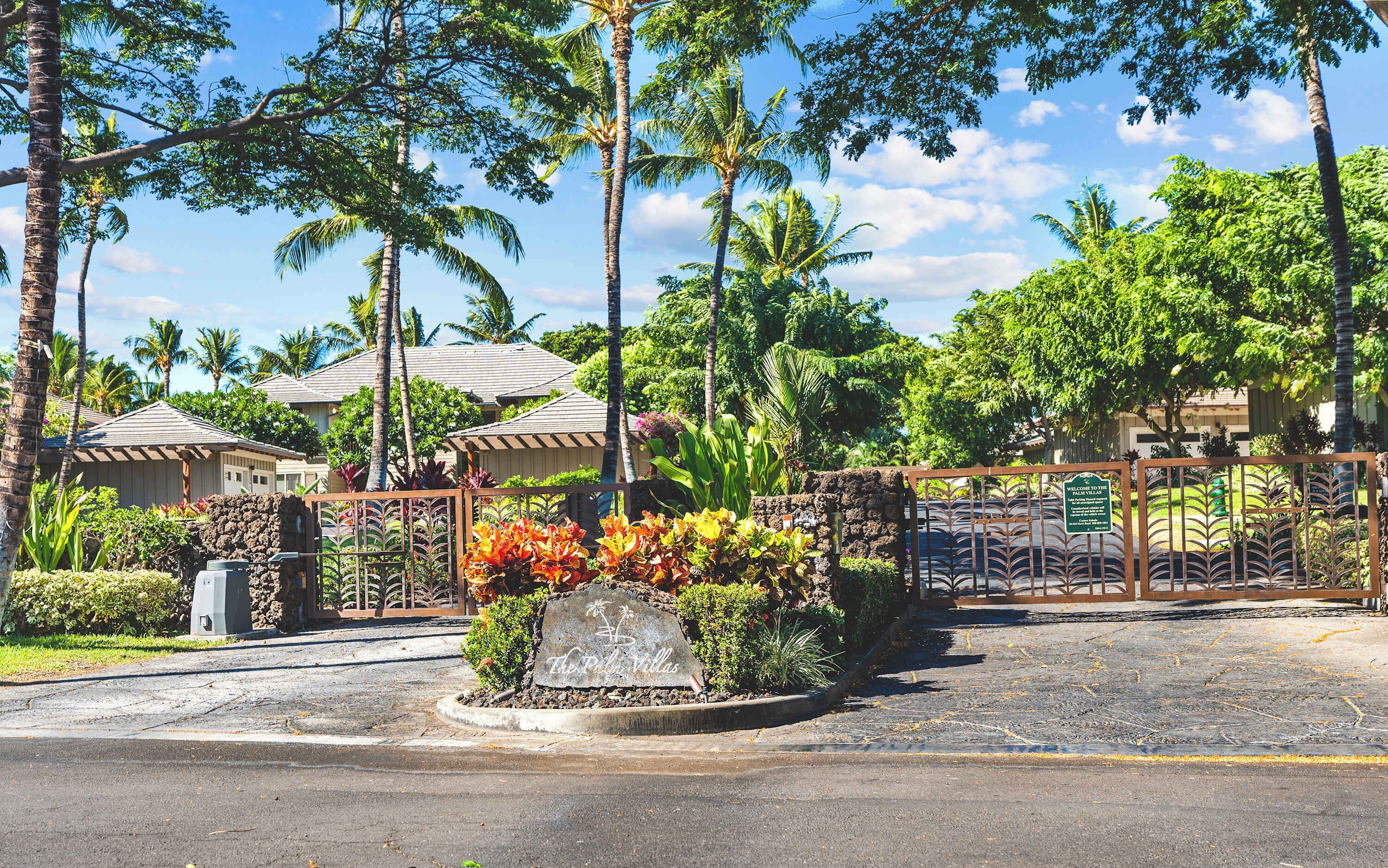 A gated entrance with a sign welcoming to the Palm Villas, surrounded by lush tropical vegetation and palm trees.