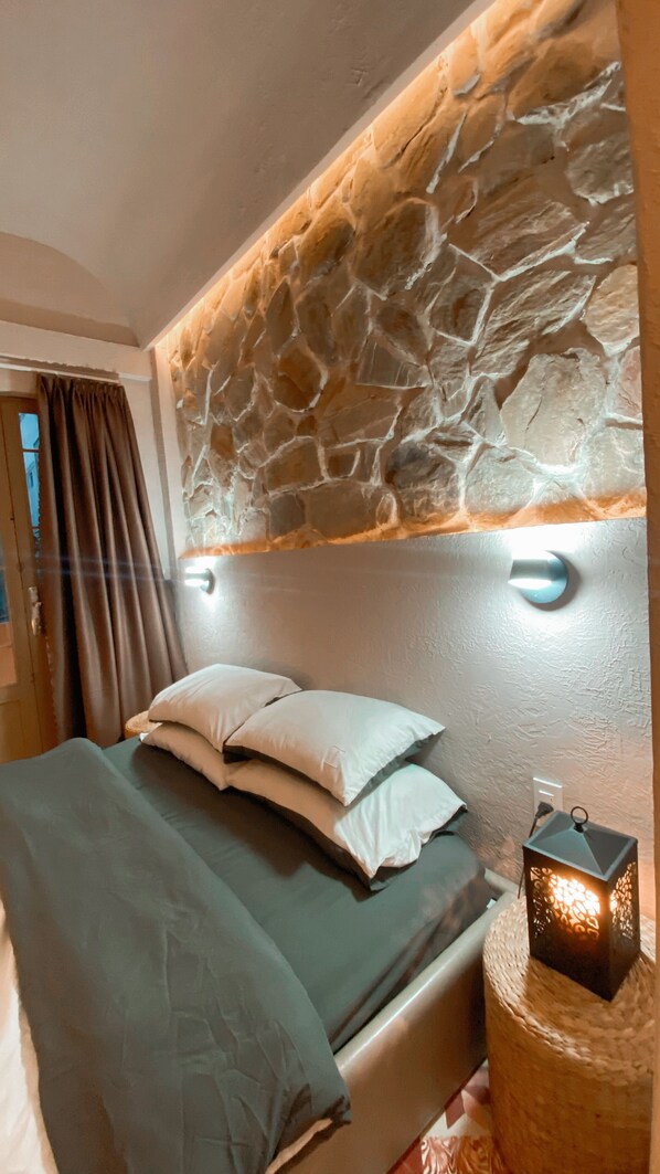 Un dormitorio con cabecera de piedra, una cama con almohadas y una mesita de noche con una linterna.
