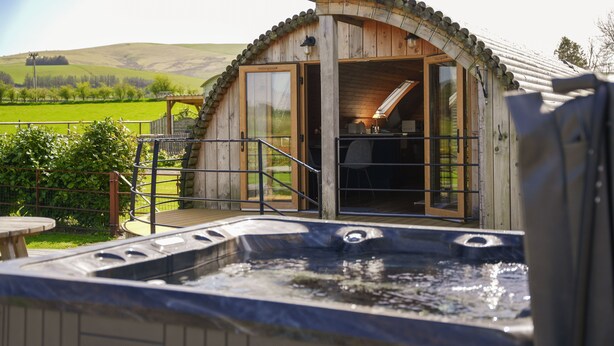 Uma cabana de madeira com banheira de hidromassagem, um deck de madeira e vista para o campo.