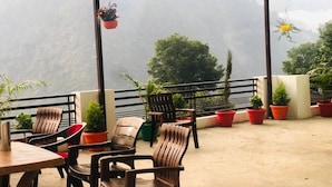 A covered outdoor seating area with chairs and tables, surrounded by potted plants, overlooking a mountainous landscape. - Hotel The Himalaya Orchid Guptkashi (Ukhimath)