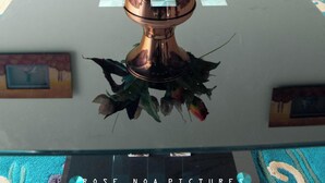A table with a copper vase containing flowers, a television in the background, and a picture frame on the table.