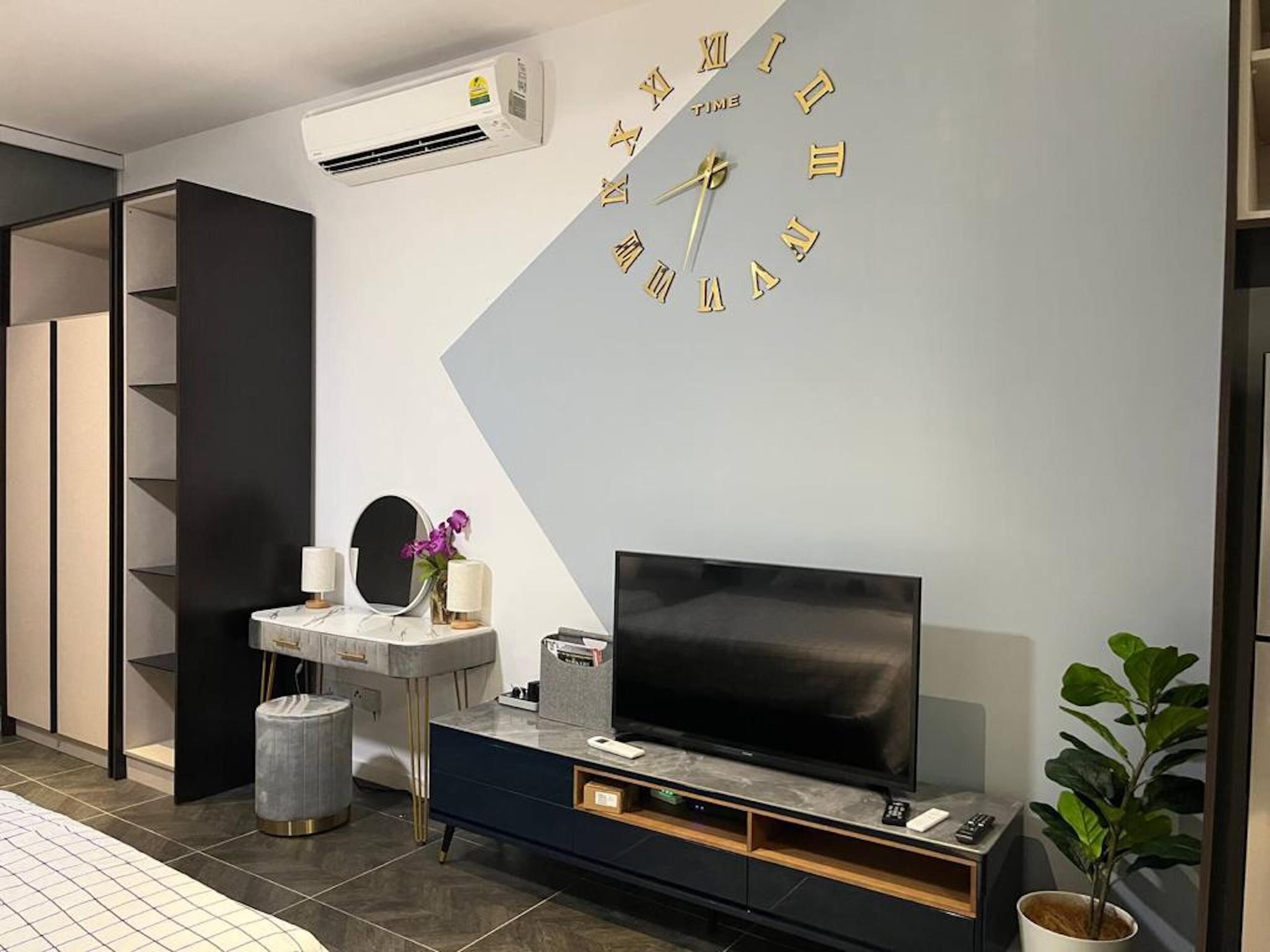 Un salón moderno con un televisor de pantalla plana grande sobre una consola oscura, un reloj montado en la pared y una mesita con un espejo y adornos.