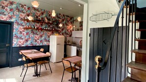 A kitchen with a vintage refrigerator, wooden dining table, and chairs. There is a staircase with a black railing and a decorative wall hanging.