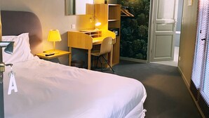A hotel room with a bed, a desk with a chair, a mirror, a lamp, and a wall-mounted shelf.
