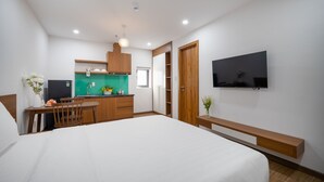 Moderni hotellihuone, jossa on suuri sänky, pieni keittiönurkkaus, seinälle asennettu televisio ja puinen penkki.