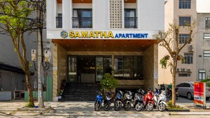 Monikerroksinen rakennus, jonka päällä on kyltti, jossa lukee ”SAMATHA APARTMENT”. Siinä on parvekkeita, joissa on kaiteet, ja sen ulkopuolella on rivi moottoripyöriä.
