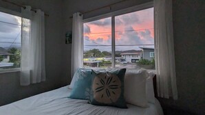 A bedroom with a bed, white curtains, and a window with a view of houses and a sunset. - Light and breezy Kailua apartment (Kailua)