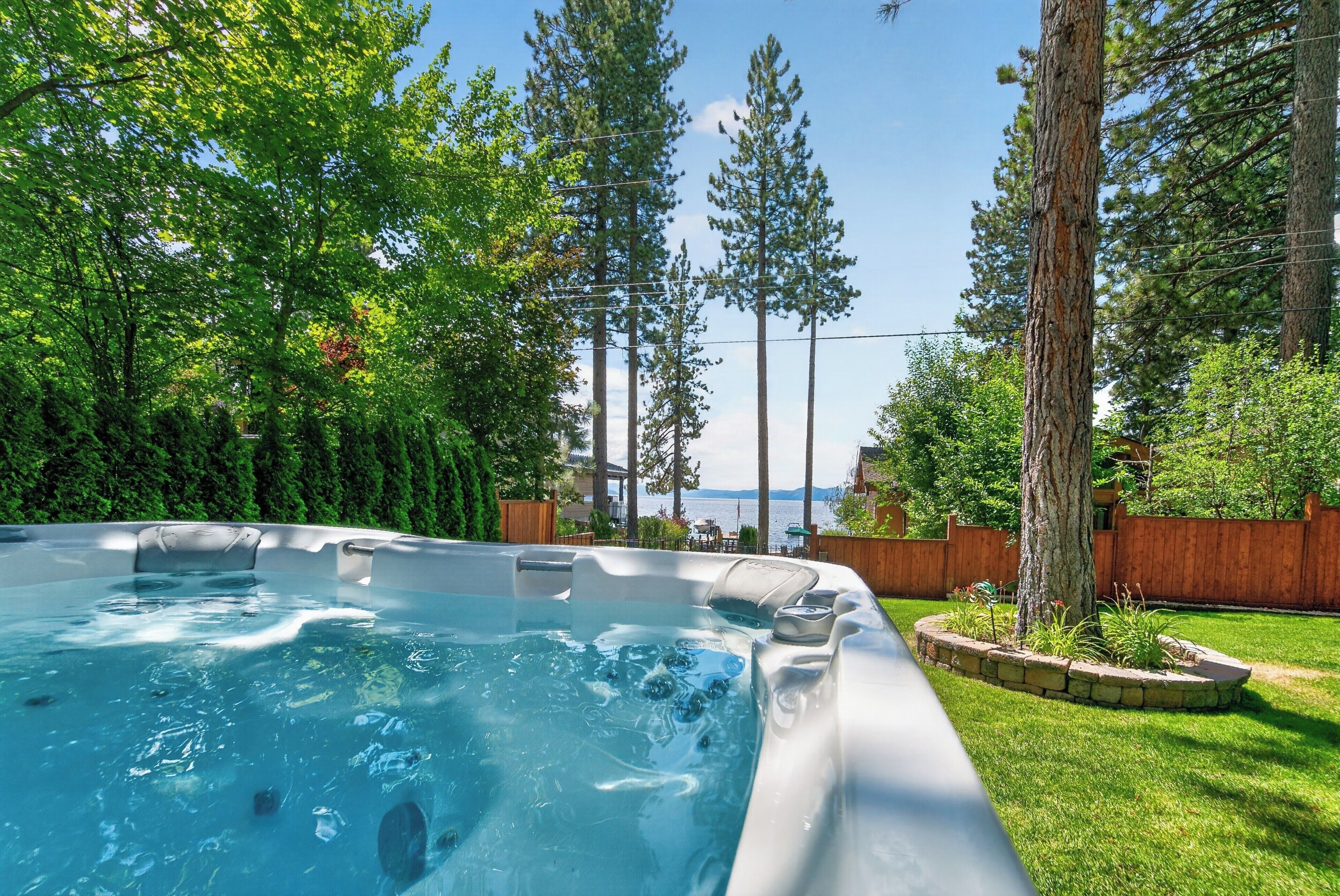 A hot tub surrounded by a wooden deck and lush greenery, with a view of a body of water and mountains in the background.