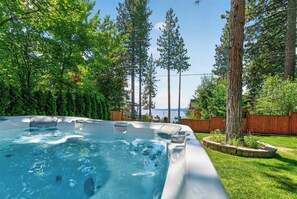 A hot tub surrounded by a wooden deck and lush greenery, with a view of a body of water and mountains in the background.